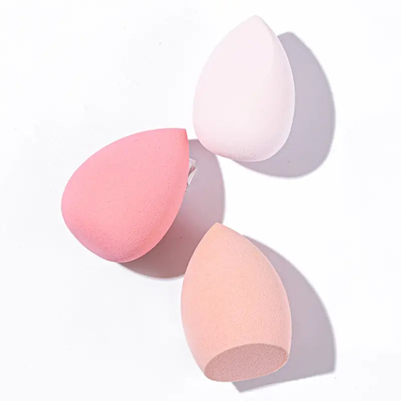 Three makeup sponges in pink, white, and peach on a light gray background