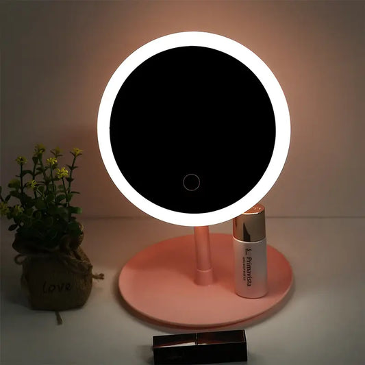 Round makeup mirror with LED lights on a pink stand, next to a plant and cosmetic bottle.